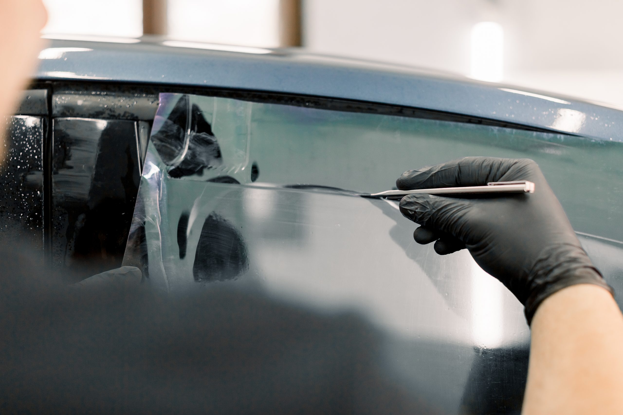 Cropped image of hands of worker in garage tinting a car window with tinted foil or film, holding special blade or knife to cut the film. Car detailing workshop, tinting windows.
